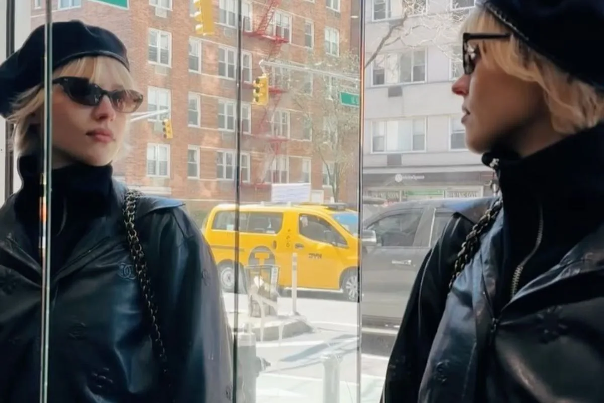 Angèle posant avec un béret noir et un manteau en cuir, reflétée dans une vitre de boutique. Elle donne un coup de pouce à la mode française grâce à son béret !