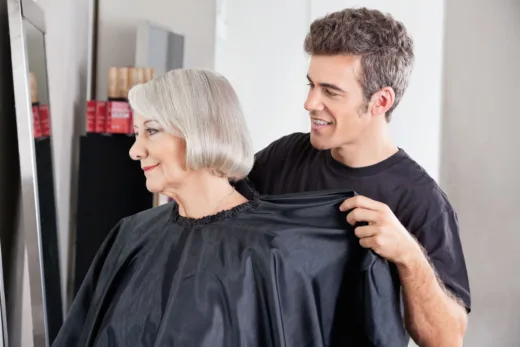 Coiffeur souriant ajustant une cape sur une cliente senior avant une coupe rajeunissante.