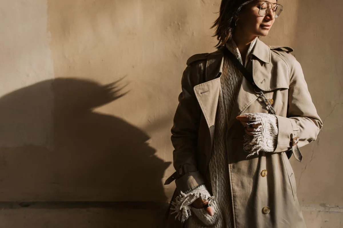 Femme élégante portant un trench-coat beige, éclairée par une lumière naturelle créant une ombre artistique sur le mur.