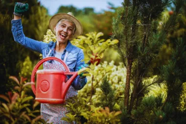 Femme âgée joyeuse avec un chapeau de paille arrosant ses plantes dans un jardin luxuriant.