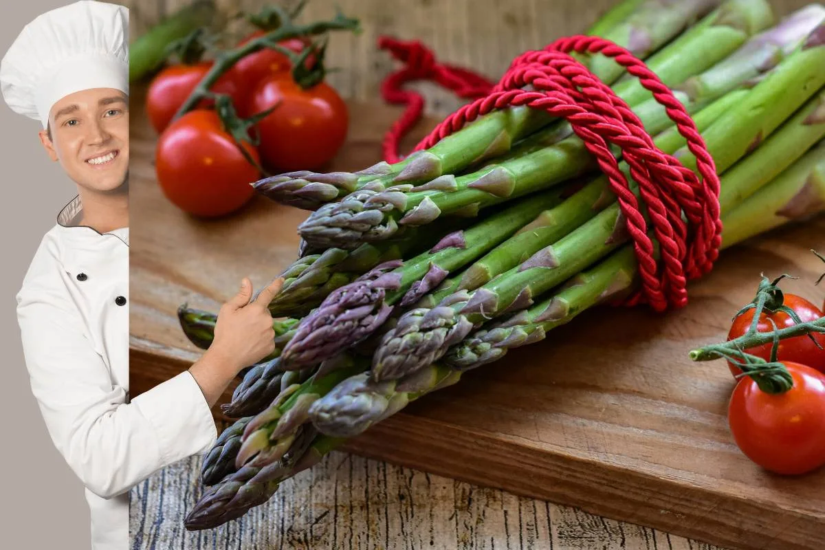 Chef souriant montrant un lien de asperges vertes et violettes fraîches sur une planche à côté de tomates rouges.